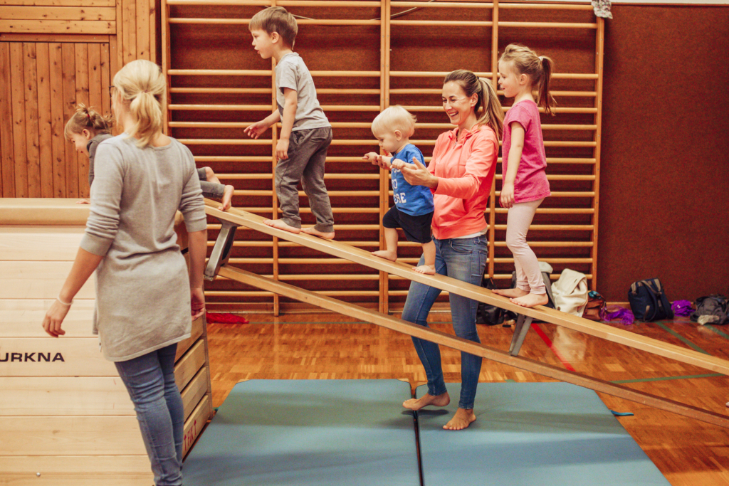 Kinder klettern mit Hilfe Ihrer Eltern auf eine schräg gestellte Langbank.